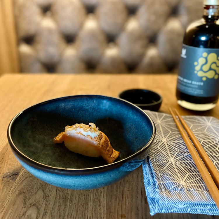 Teller mit Nigiri Sushi. Daneben ein Tuch mit Esstäbchen und dahinter ein Sojasaucenschälchen und eine Flasche mit Sojasauce.
