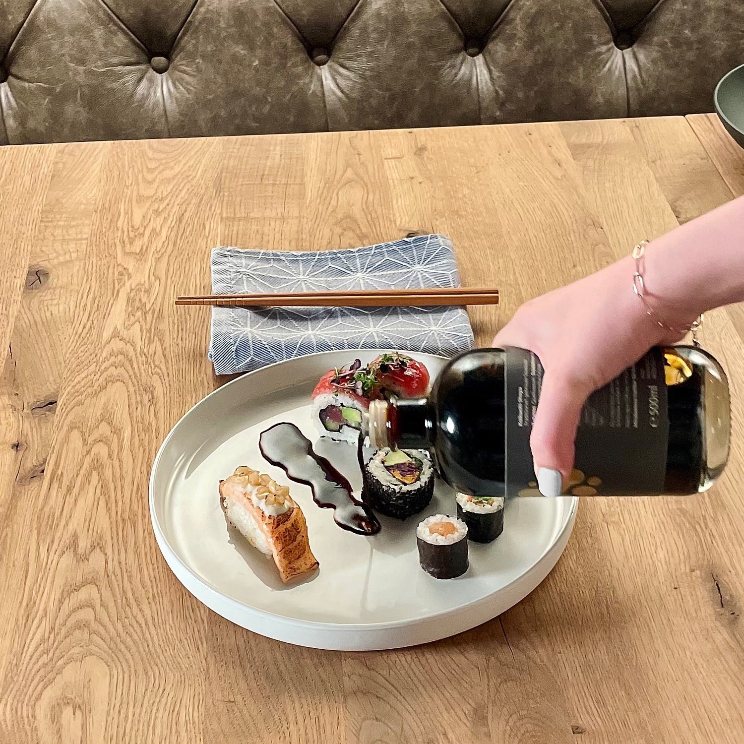 Holztisch mit weißem Teller mit Sushi Variationen. Daneben Tuch mit Essstäbchen. Eine Hand gießt aus einer Flasche Sojasauce auf den Teller.